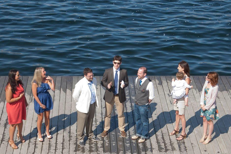 Drew Miller and Kyle Miller face each other on a floating dock with an officiant between them and two witnesses each on the left and right. The boards of the deck are wet in the center and edges.