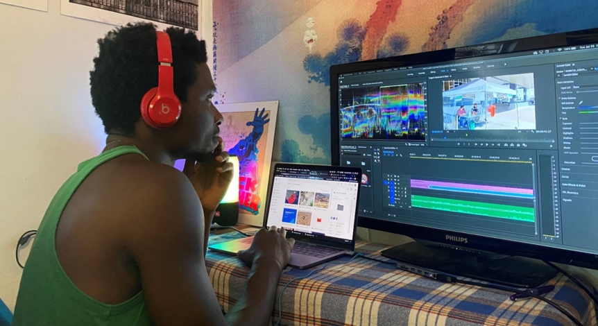 A student in a green tank top and red headphones is sitting in front of three brightly colored computer monitors editing a video.