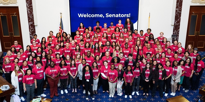 A group of approximately fifty people smiling in matching red shirts in front of a banner that says "Welcome, Senators." 