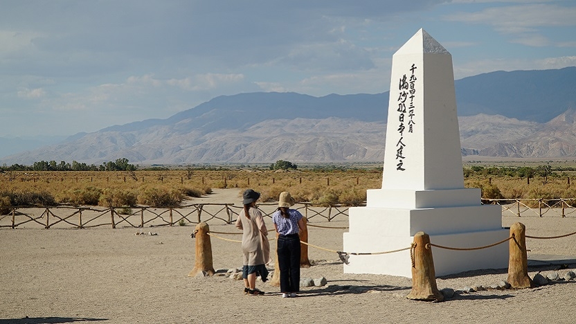 Two people stand in front of a white monument with Japanese lettering set behind a fence. In the distance, you can see mountains and blue sky.