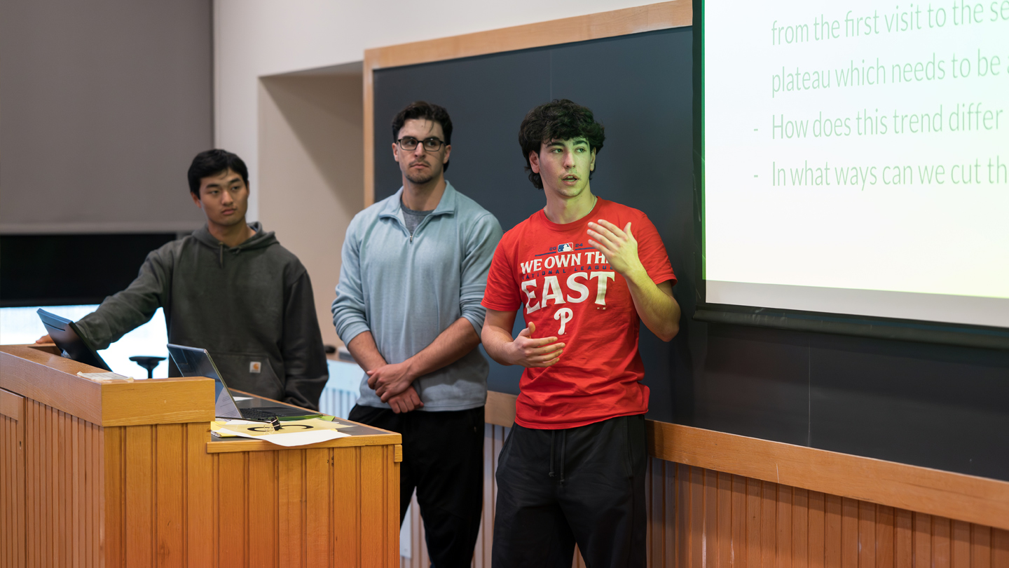 Three students stand at a podium giving a presentation in a lecture hall.