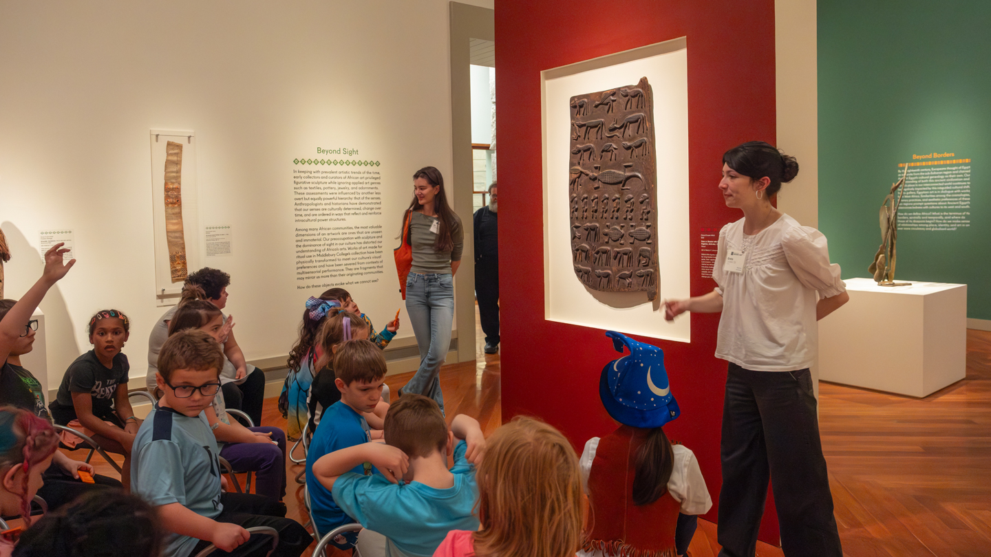 Two college students stand beside a wooden carved door hanging on the wall of the art museum, and explain it to a group of school children.