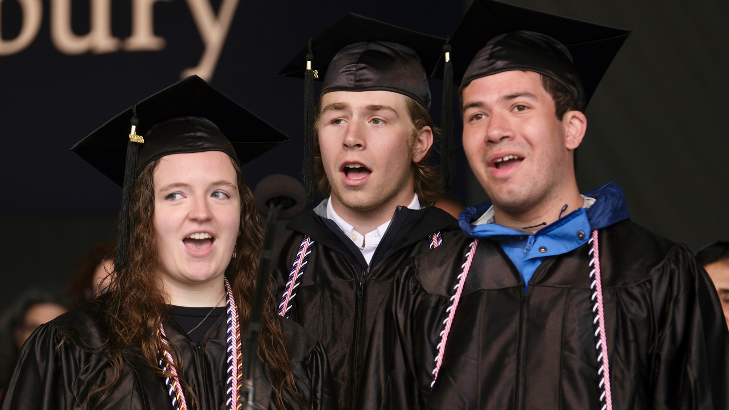 Three students in graduation caps and gowns sing the national anthem on a stage.