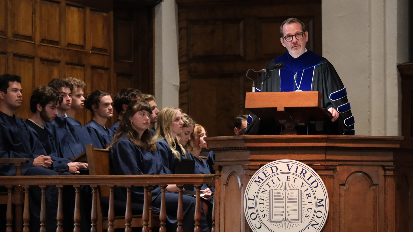 President Ian Baucom in academic regalia stands at a podium in Middlebury Chapel
