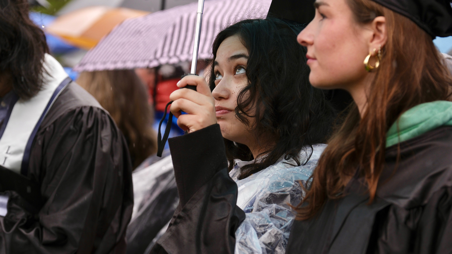 Students in commencement robes stand under umbrellas.