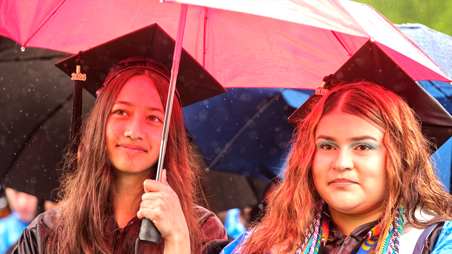 Two students at graduation under a red umbrella.
