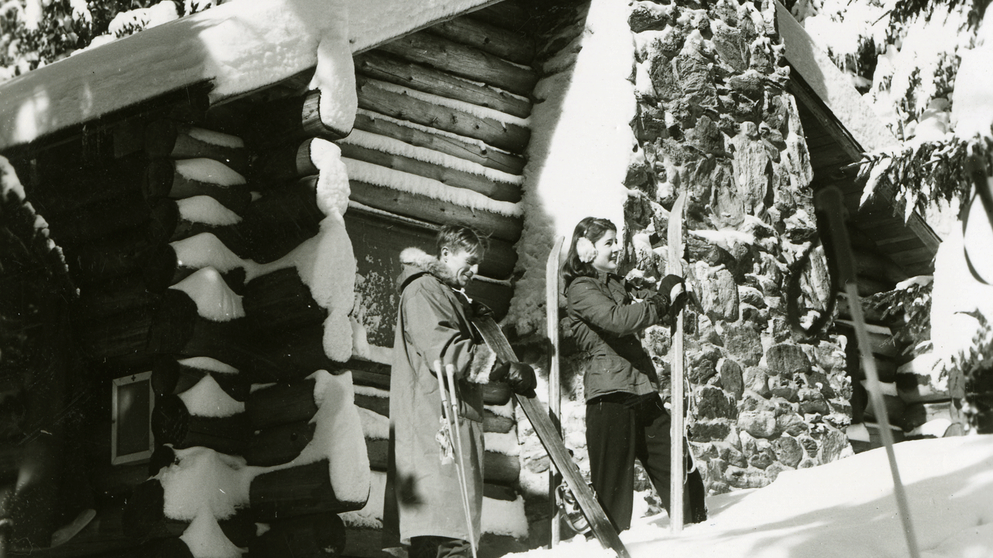 A historic black and white photo of the Hubbard log cabin from the 1950s with two students with skis in front of the door.