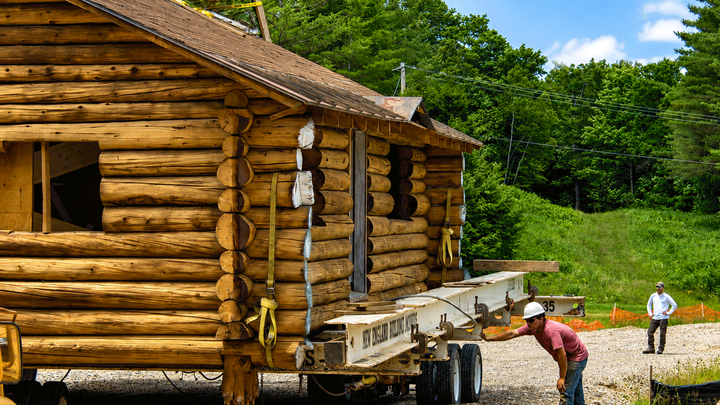A worker wearing a hardhat and orange shirt checks the Hubbard log cabin as it is being towed to a new location at the Middlebury College Snowbowl.