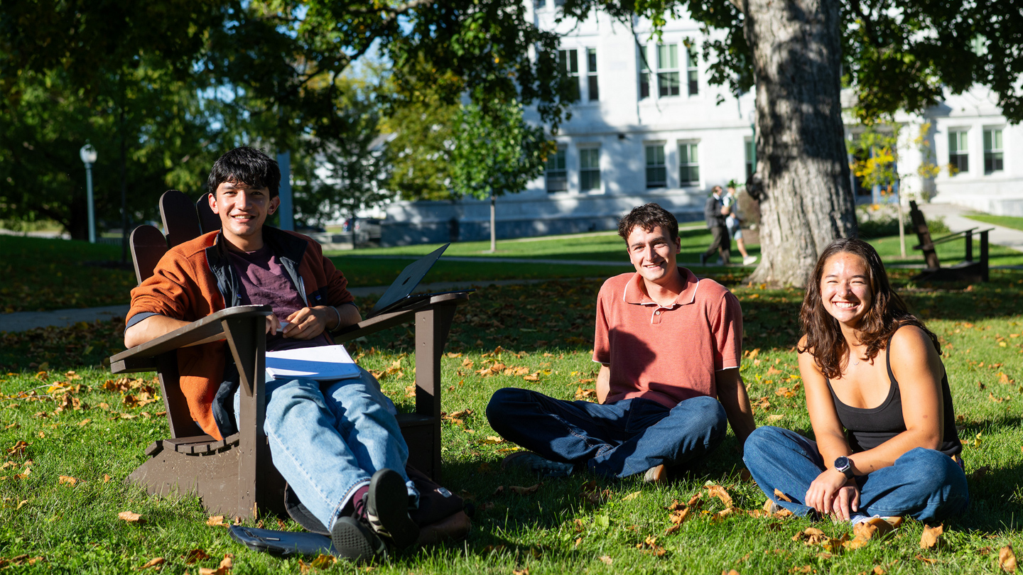 Three students sit on the lawn in front of a campus building, one in an Adirondack chair.