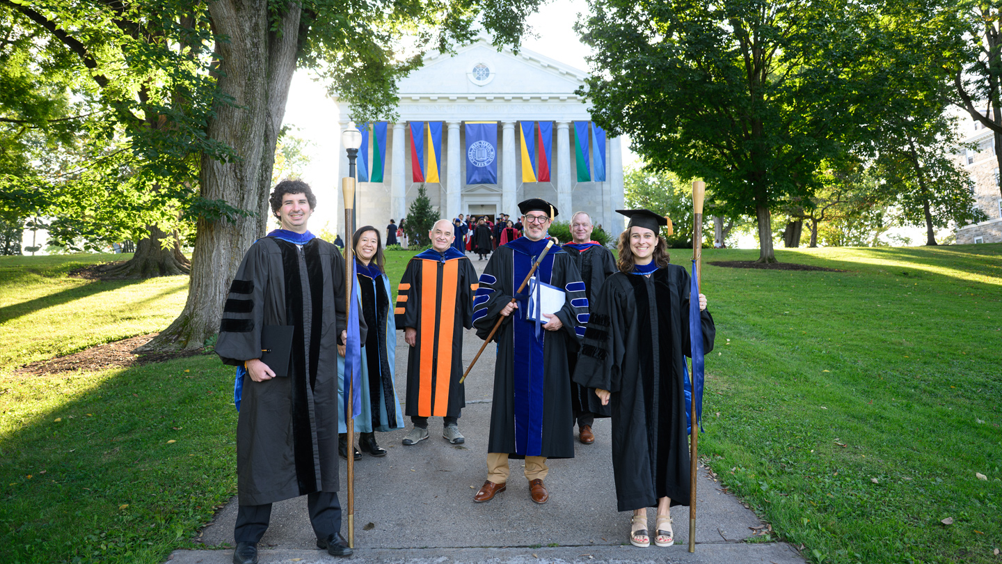 Professors and administrators in academic regalia smile for a group photo outside Middlebury Chapel.