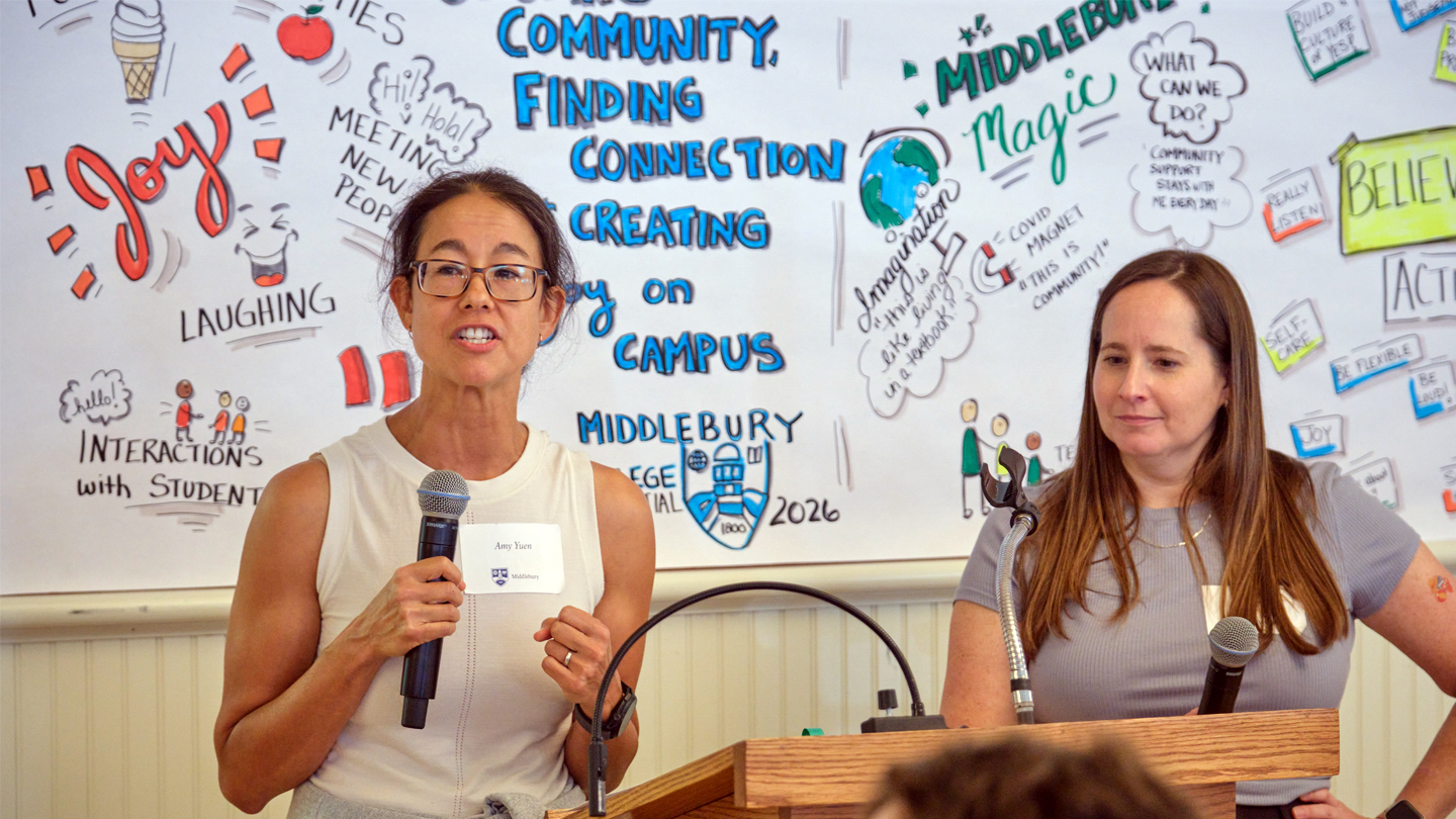 Two people stand in front of an illustrated mural, one speaking into a microphone.