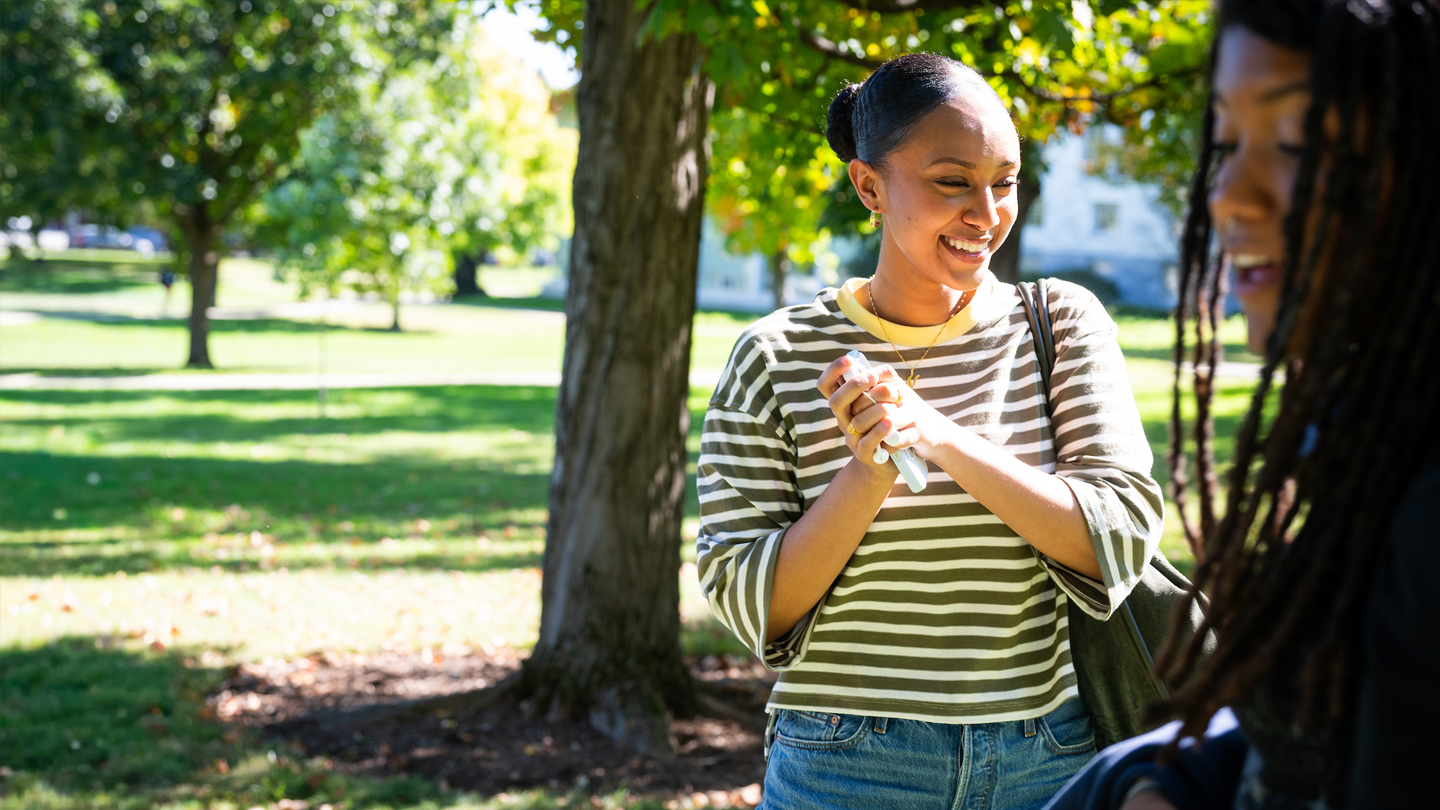 A smiling student in a striped top chats with another person under a tree.