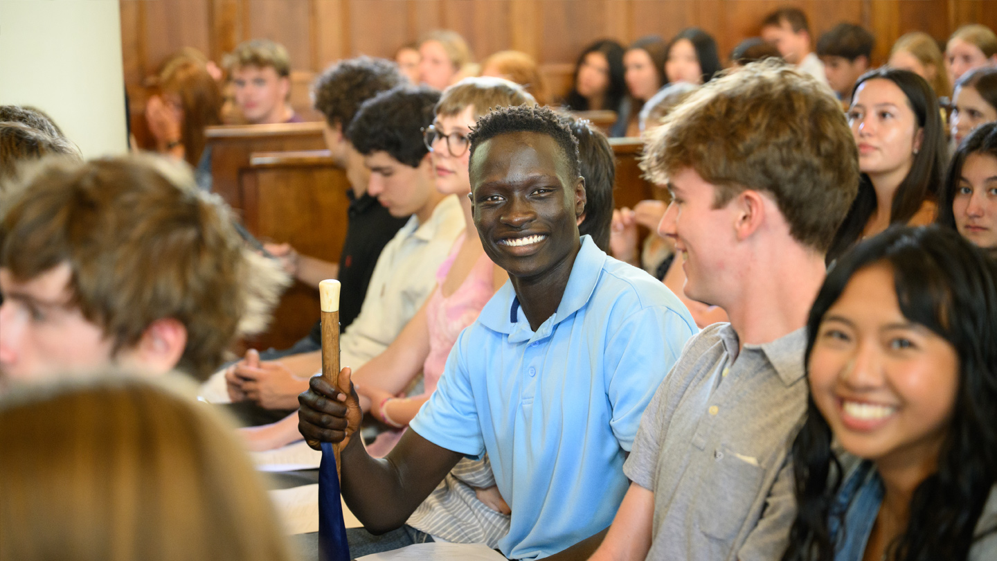 A student wearing a light blue shirt sits in a pew with other students and smiles at camera while holding a cane.