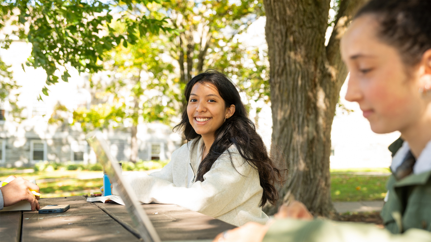 A student smiles at the camera while studying outside at a picnic table.