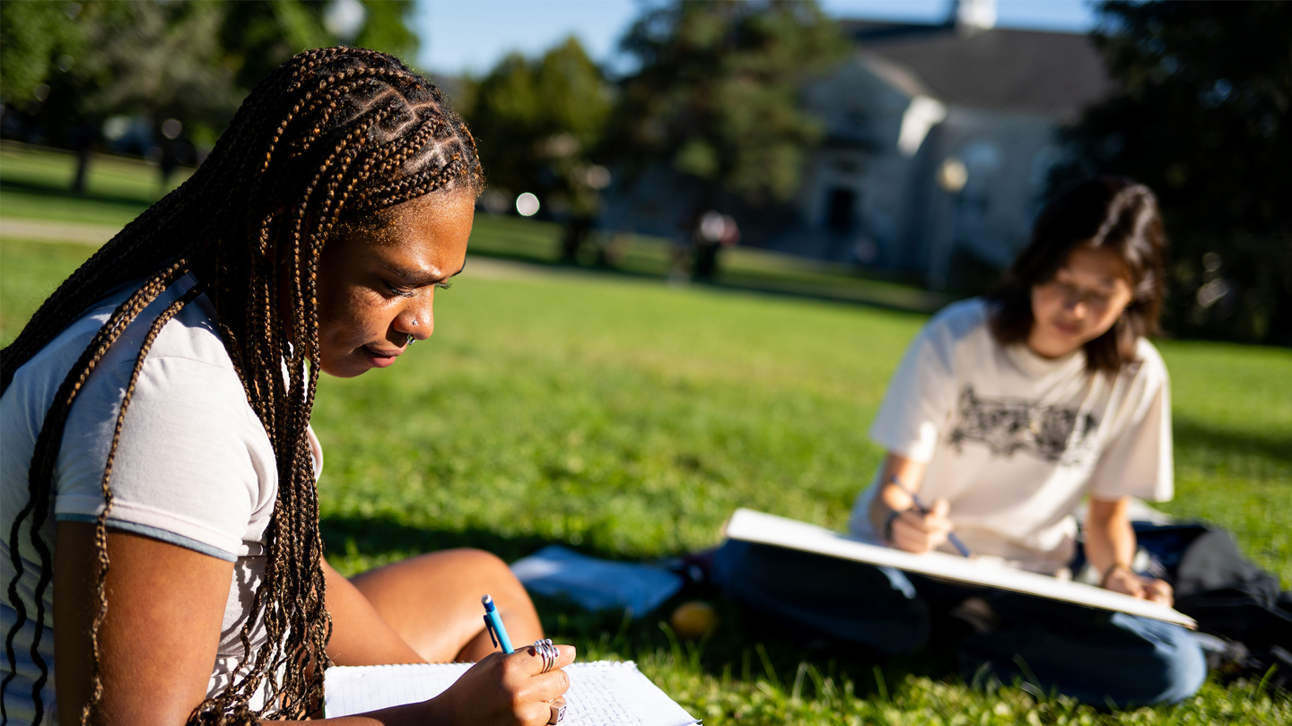 Two students sit on a campus lawn writing in notebooks.