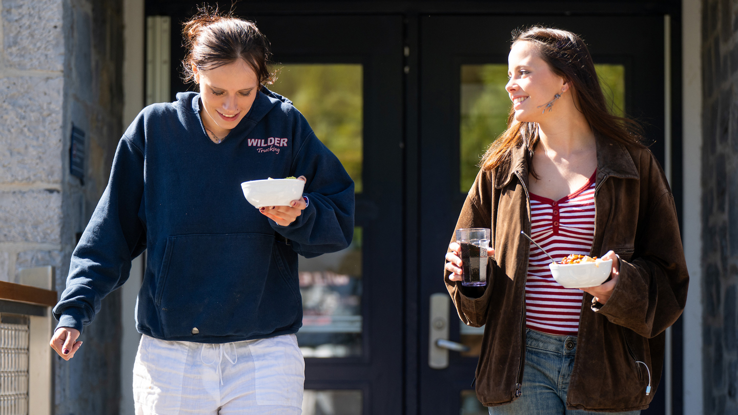 Two students walk out of a building carrying bowls of food.