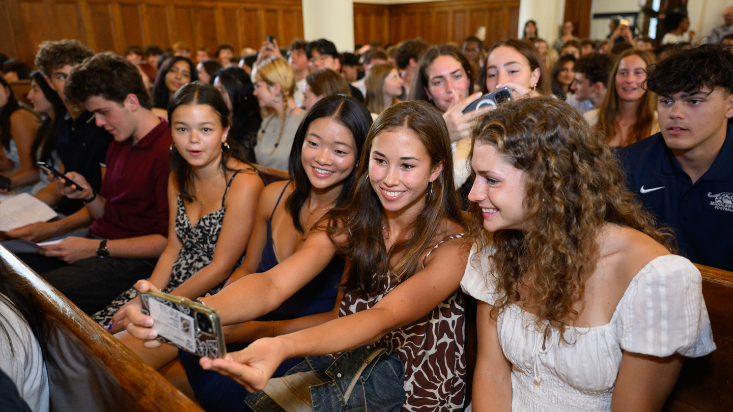 Students sitting in a pew take a selfie surrounded by classmates.