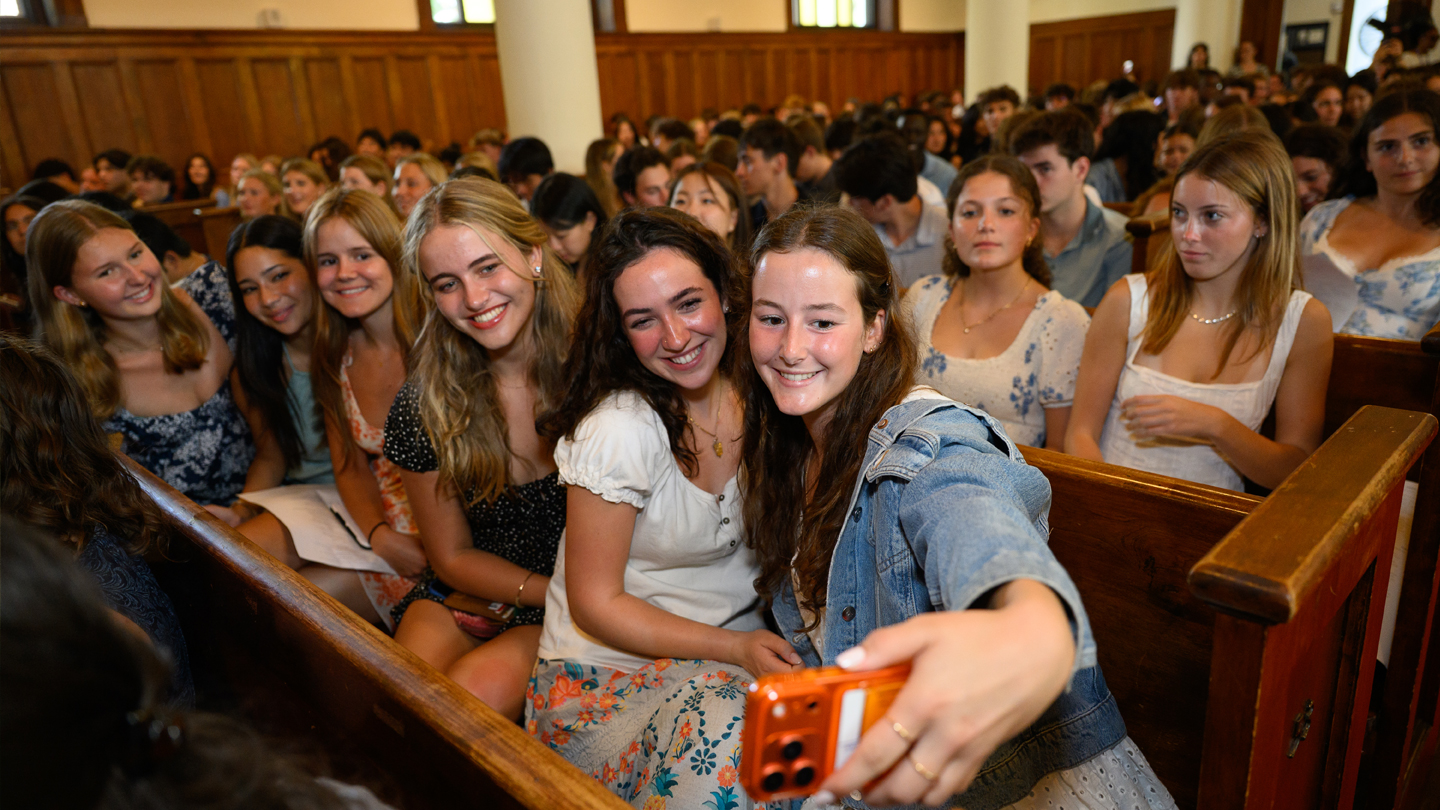 Students in chapel pews take a group selfie surrounded by smiling classmates.
