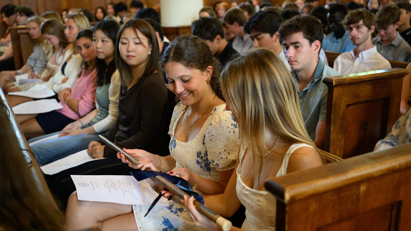 Students sitting in a pew look at a cane as they pass it from one person to the next.