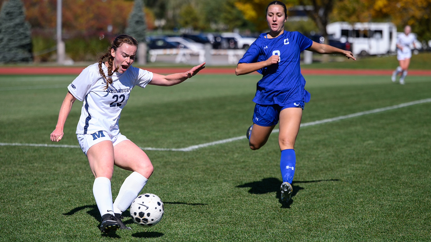A woman in a Middlebury uniform kicks a soccer ball. An opposing player is nearby. 