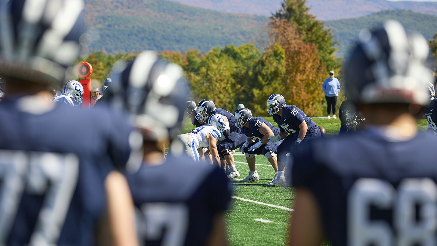 The foreground of the photo is blurry, looking through a group of football players on the sidelines. The background is in focus, and you see a formation of football players on the field ready to snap into action. 