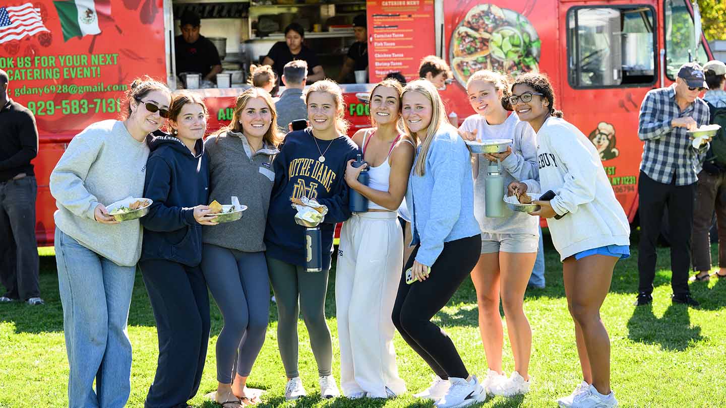 A group of students poses for a photo in front of a food truck. They are holding plates of tacos. 