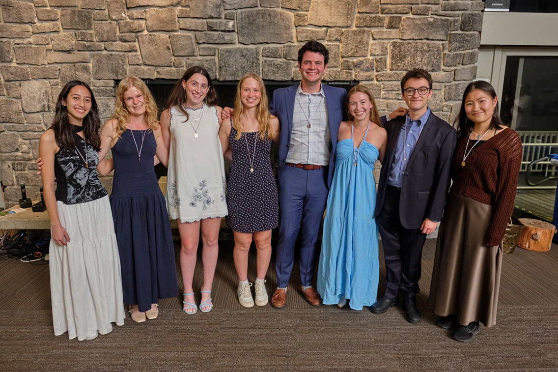 A group of students stands in a line in front of a field stone fireplace.