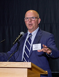 Man in dark blue suit and striped tie speaks at podium.