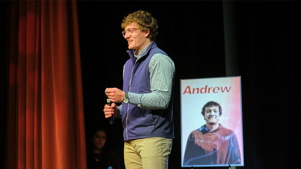 A student wearing a blue vest speaks into a microphone on a stage.