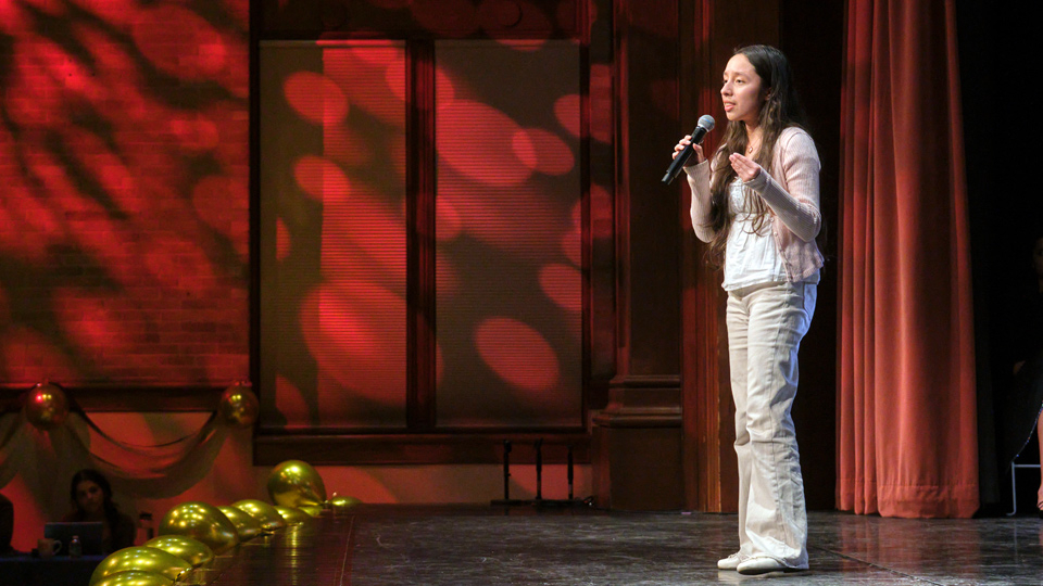 A student wearing white stands on a stage with a microphone.