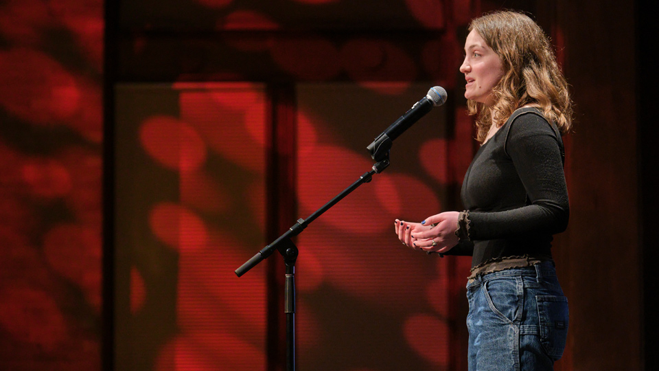 A student wearing a dark sweater speaks on a stage.