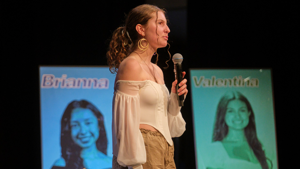 A student wearing a white top speaks into a microphone on stage.
