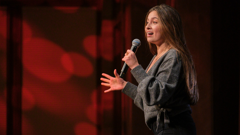 A student wearing a sweater speaks into a microphone on a stage.