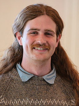 Student wearing a blue shirt with brown sweater smiles at the camera.