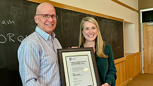 Two professors hold a framed award and smile at the camera during a ceremony.