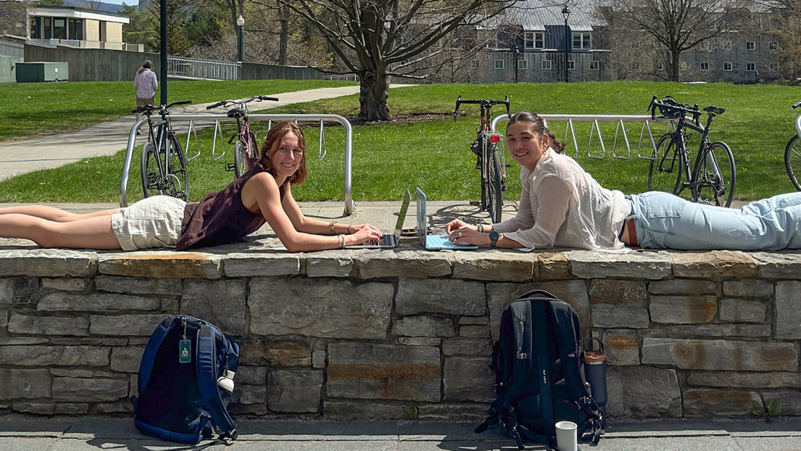 Two students on a stonewall on campus with laptops.