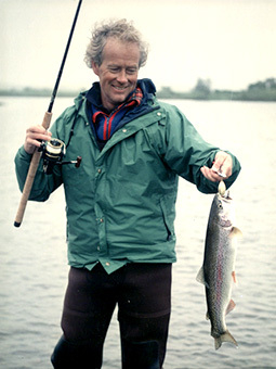 Dort Cameron stands in a body of water wearing fishing gear and holding a fish.