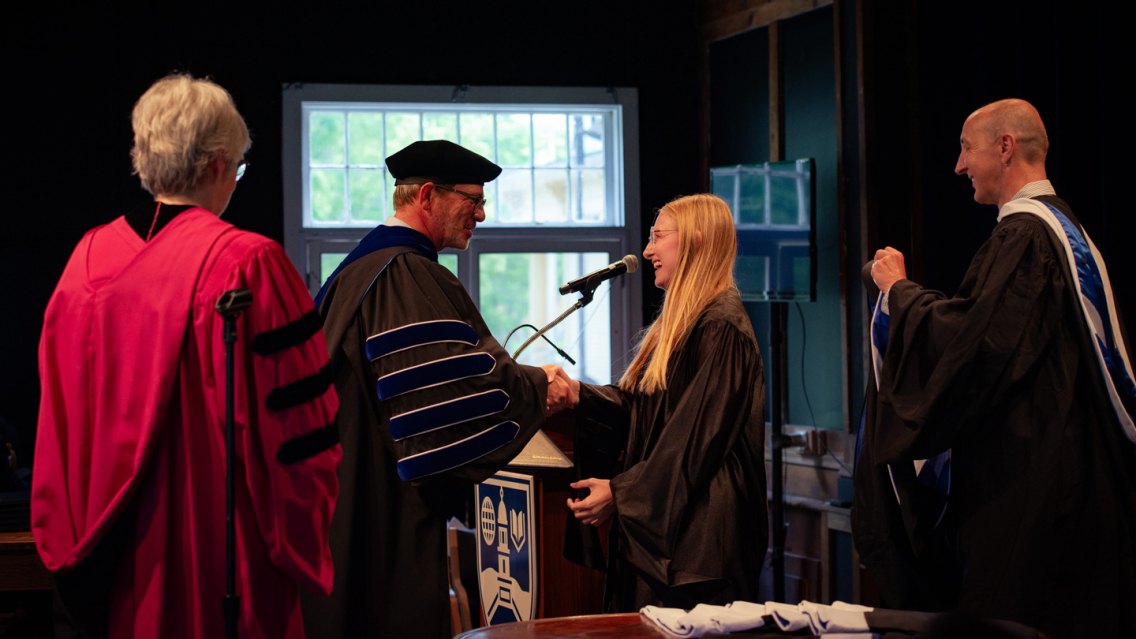 In academic regalia, Middlebury President Ian Baucom shakes hands with a new graduate of the Bread Loaf School of English as she crosses the stage.