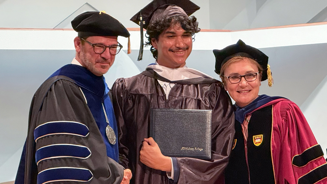 President Ian Baucom stands with a student holding their diploma and a director  