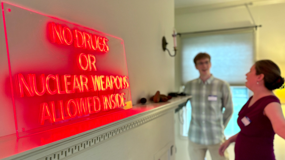 A glowing neon red sign that says "no drugs or nuclear weapons allowed" hangs above a mantlepiece in a house.