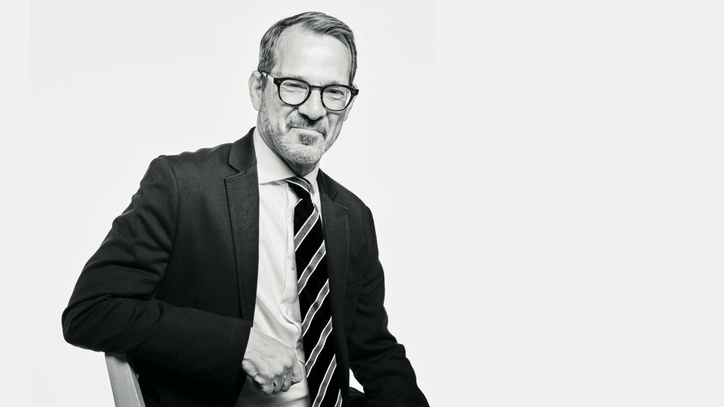 A black and white photo of President Ian Baucom wearing a suit and smiling at the camera.