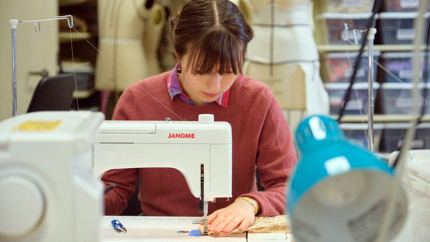 A student wearing a red sweater works at a sewing machine.