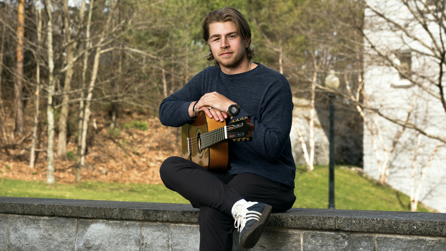 A Middlebury student sits outside on a stone wall with a guitar.