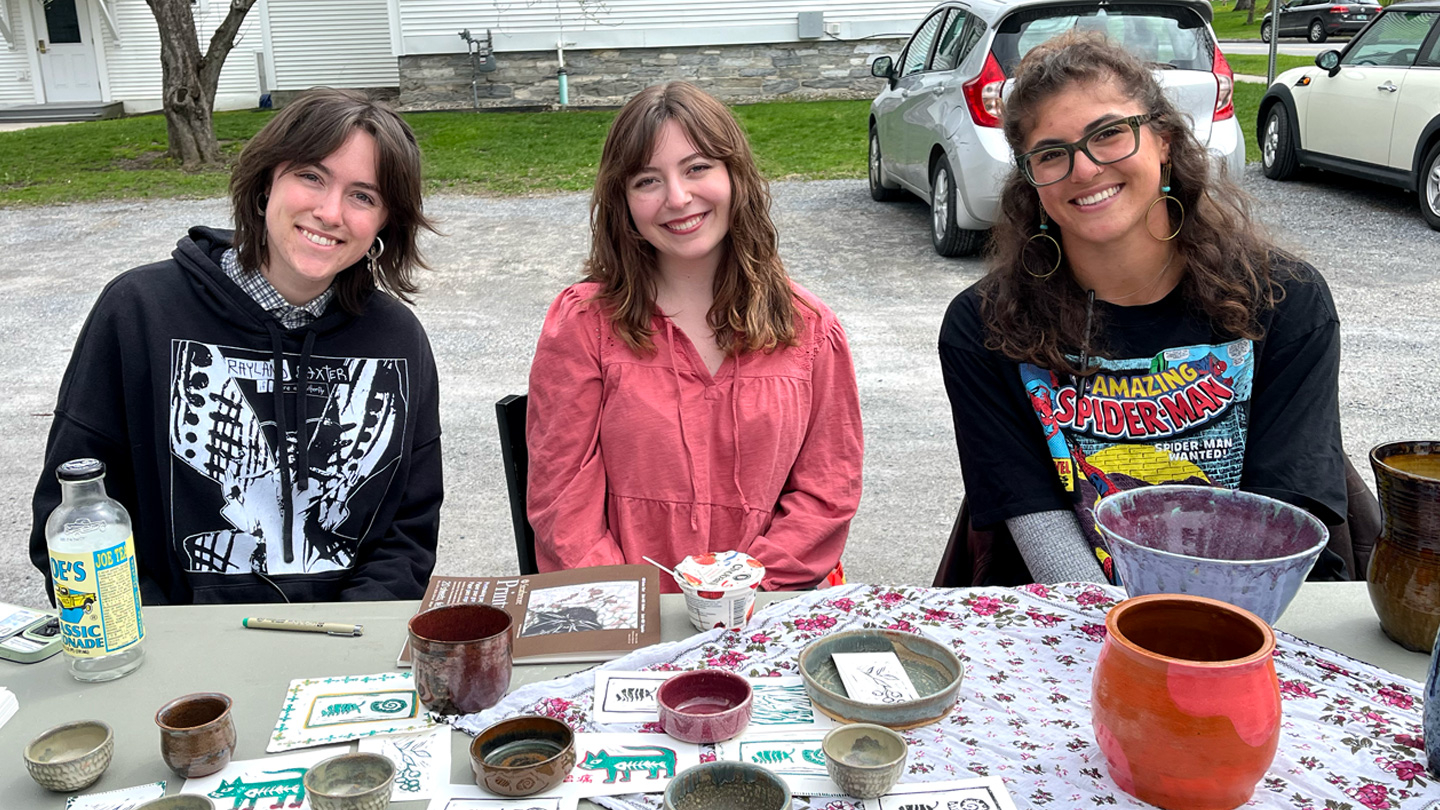 Three students sit at a table outside with ceramic art for sale