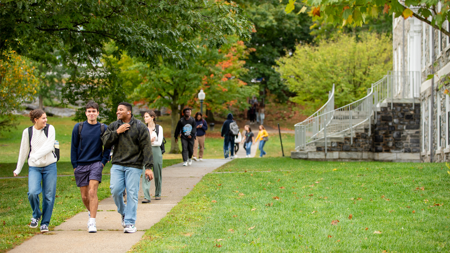 Students walk along a sidewalk on a leafy college campus.