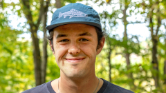 A student with a blue baseball hat smiles at the camera while standing in the woods.