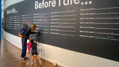 A parent and child stand at a large blackboard in a hallway.