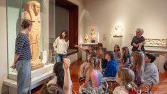 Two college students discuss a sarcophagus in the museum with school children.