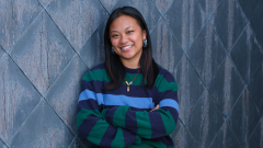 A student in a dark sweater leans up against a patterned wall outside.