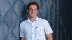 A student in a white shortsleeved shirt leans against a patterned wall outside.